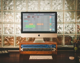 May include: A desktop computer setup featuring a large monitor displaying audio editing software. Below the monitor is a wooden rack with audio equipment. A keyboard, mouse, and other audio gear are on the desk. The background includes a glass block wall.