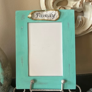May include: A teal blue picture frame with a white mat and a gold and white decorative plaque that says "Family". The frame is on a white metal stand and a string of blue beads.
