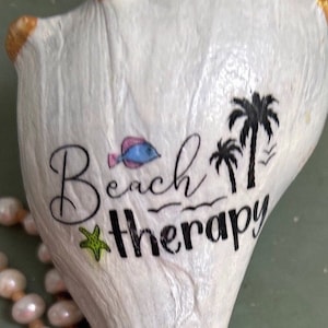 May include: White seashell with gold accents, painted with the words "Beach Therapy" and a palm tree design. A small blue fish and a green starfish are also painted on the shell.