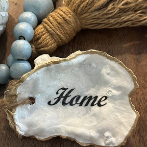 May include: A white oyster shell with a gold rim, painted with the word "Home" in black script. The shell is attached to a string of light blue wooden beads and a brown jute tassel.