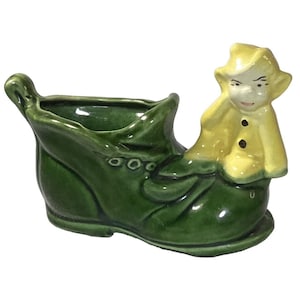 Peut inclure: Un pot de fleurs en céramique en forme de botte verte avec une poignée. Une figurine d'une personne en manteau et chapeau jaunes est assise sur la botte. Le pot a trois petits cercles sur le côté et est sur fond blanc.