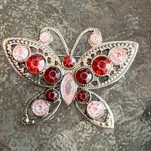 May include: A silver-toned butterfly brooch with a detailed filigree design. The brooch features a symmetrical design with red and pink gemstones of various sizes, creating a sparkling effect. The butterfly's body is a light pink oval gem.