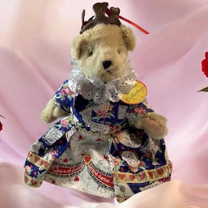 May include: A plush teddy bear dressed as the Queen of Hearts, wearing a blue and white floral dress with red heart accents. The bear has a brown crown and a ruffled collar. A tag reads "Collection".