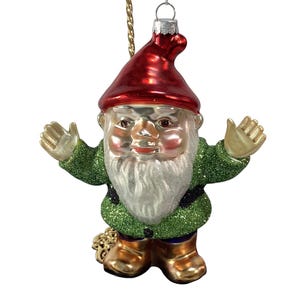 Pode incluir: Enfeite de Natal de vidro em forma de gnomo de jardim. O gnomo tem um chapéu vermelho, barba branca e casaco verde com glitter dourado. O enfeite é suspenso por uma corrente dourada e um fio prateado.
