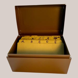 May include: A brown metal card file box with the lid open, showing yellow index cards. The cards are labelled with black handwritten letters A to E, indicating an alphabetical filing system. The box is rectangular and has a hinged lid, typical of a filing system.