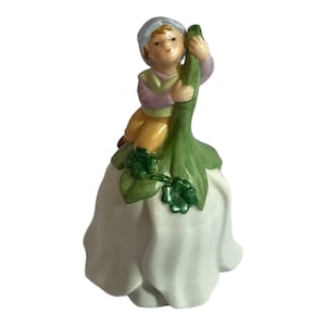 May include: A ceramic figurine of a small child in a blue cap and colorful clothing, perched on a large white flower. The flower has green leaves and a green stem. The figurine is a decorative item.