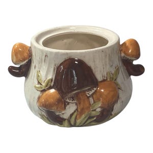 Puede incluir: Un recipiente de cerámica con tapa, con un diseño de setas. El recipiente es de color blanquecino con setas marrones y naranjas y hojas verdes. Dos asas con forma de seta están a cada lado.