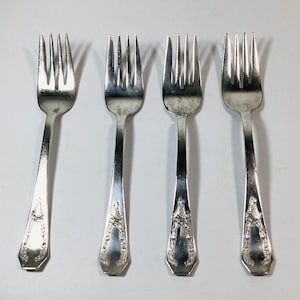 Puede incluir: Cuatro tenedores plateados con púas y mangos decorativos. Los tenedores están dispuestos sobre una superficie blanca. Cada tenedor tiene un diseño floral detallado en el mango. Los tenedores son de metal brillante y reflectante.