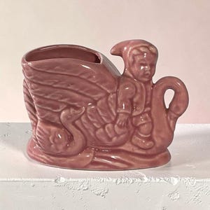 Könnte beinhalten: Ein vintage-rosa Keramik-Blumentopf in Form eines Schwans, der eine kleine Figur trägt. Der Schwan hat detaillierte Flügel und einen gebogenen Hals, wobei die Figur auf seinem Rücken sitzt. Der Topf hat eine glatte, glänzende Oberfläche.