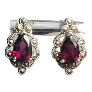 May include: A pair of silver-toned stud earrings. Each earring features a teardrop-shaped, deep red gemstone surrounded by small, round, silver-toned accents. The earrings have a vintage or antique aesthetic.