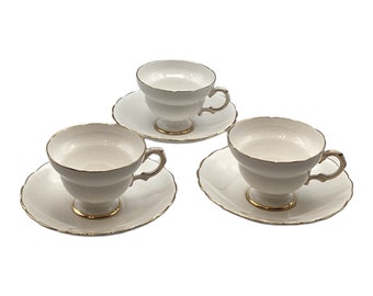 May include: Three white teacups and saucers with gold trim. The teacups have a pedestal base and curved handles. The saucers have a scalloped edge. The set is displayed on a white background.