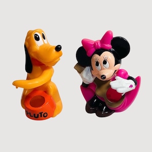 Könnte beinhalten: Zwei Plastikfiguren von Disney-Charakteren. Pluto, ein gelber Hund, sitzt mit einem orangefarbenen Behälter mit der Aufschrift "PLUTO". Minnie Mouse trägt ein rosa Kleid und eine Schleife, weiße Handschuhe und schwarze Ohren.