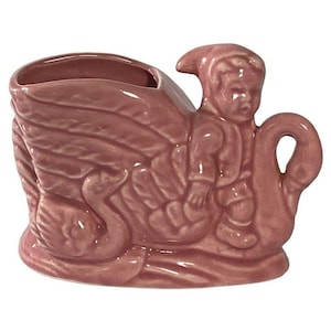 Könnte beinhalten: Ein Vintage-Blumentopf aus rosa Keramik in Form eines Schwans mit einer kleinen Figur, die auf seinem Rücken reitet. Der Pflanzentopf hat ein detailliertes Design von Federn und der Schwanenhals bildet einen Griff. Die Figur trägt einen Hut und einen Anzug.