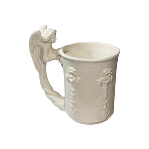 May include: A cream-colored ceramic mug with an angel-shaped handle. The handle features a detailed angel figure with outstretched wings. The mug's body is adorned with floral relief patterns. The mug is a decorative item.