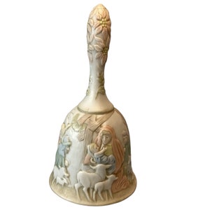 May include: Cream ceramic bell with a handle decorated with floral and leaf patterns. The bell has a relief scene of the Nativity, with figures in muted colours. A decorative item for religious or holiday decor.