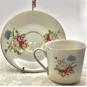 Puede incluir: Juego de taza y platillo de té color crema con diseños florales. La taza y el platillo tienen ribetes dorados y presentan un patrón de flores rosas, blancas y azules con hojas verdes. El juego se exhibe sobre un mantel de encaje.