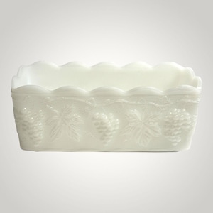 May include: A rectangular, white, milk glass dish with a scalloped rim. The sides are decorated with embossed grape clusters and leaves. The dish has a classic, vintage aesthetic.
