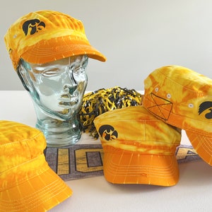 May include: Several yellow and orange tie-dye style cadet caps with a black logo. The caps are displayed with a glass head and a yellow and black pom-pom. The caps have a structured brim and a stitched design.