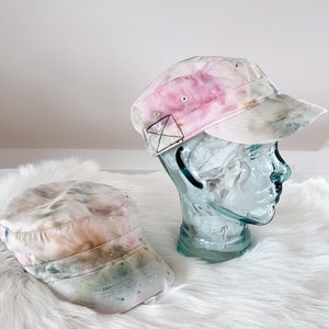 May include: Two tie-dye army caps, one pink and white, the other green and white, on a white fluffy surface.