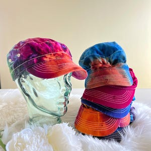 May include: A stack of colorful tie-dye visors and a tie-dye cap with a curved brim. The cap is displayed on a glass head. The visors and cap feature a mix of colors, including pink, orange, blue, and purple. White stitching accents the brims.