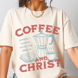 May include: A beige t-shirt with a graphic design of a coffee pot, a cup of coffee, and an open book. The text on the shirt reads "Coffee and Christ" with the words "When I wake give me Jesus" written in a smaller font.