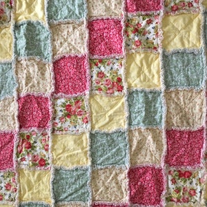 May include: A patchwork quilt featuring a range of floral designs in shades of pink, yellow, and green. The quilt is made with a simple square pattern and has a frayed edge.