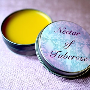 May include: A small, round tin container with a yellow balm inside. The lid of the tin has a blue and pink floral design with the text "Nectar of Tuberose" in purple.