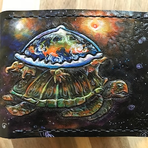 Op de afbeelding: Een zwarte leren portemonnee met een kleurrijk schilderij van een schildpad die de aarde op zijn rug draagt. De achtergrond is donkerblauw met sterren en een feloranje zon.