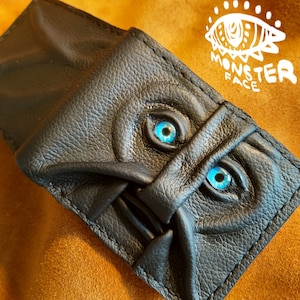 May include: Black leather wallet with a stitched design resembling a monster face with two blue eyes. The wallet has the text "Monster Face" printed on it.