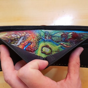 Puede incluir: Una cartera de cuero negro con una escena pintada a mano colorida en el interior. La escena representa un paisaje con un castillo, árboles y un río.