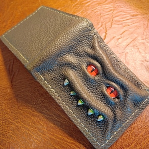 May include: A brown leather wallet with a textured surface. The wallet features a design of a creature's face, with two red eyes and several small, pointed teeth. The stitching is a light beige color.