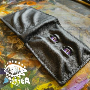 May include: A black leather wallet with two purple eyes sewn on the front. The wallet is folded closed and lying on a colorful surface.