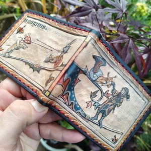 Puede incluir: Una cartera de cuero beige con ilustraciones medievales grabadas y pintadas a mano que muestran conejos en combate y una figura con un hacha. La cartera tiene bordes negros y costuras rojas a lo largo de todo el perímetro.