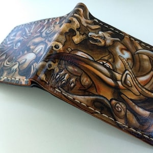 May include: A brown leather wallet with a detailed hand-painted design of a dragon and other mythical creatures. The wallet is closed and the design is visible on both sides.