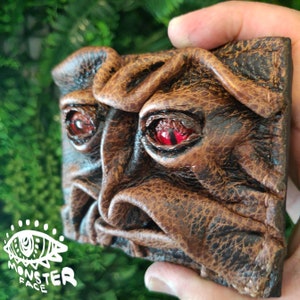 May include: A brown leather wallet with a monster face design. The wallet has two red eyes and a textured surface.