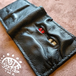 May include: A black leather wallet with a unique design featuring two eyes, one red and one white, sewn into the front.