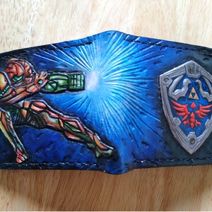 May include: A blue leather wallet featuring a painted image of a video game character, Samus Aran from Metroid, firing a beam weapon. The wallet also includes a silver and red shield with a stylised bird and a triangle design.