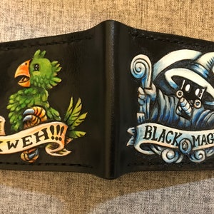 Könnte beinhalten: Schwarze Ledergeldbörse mit handgemalten Illustrationen. Eine Seite zeigt einen grünen Vogel mit dem Text "Kweh!" und die andere Seite zeigt einen blauen und weißen schwarzen Magier mit dem Text "Black Mage".