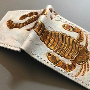Pode incluir: Uma carteira de couro branco com um design de escorpião em relevo dourado. A carteira está dobrada ao meio e tem uma borda costurada.