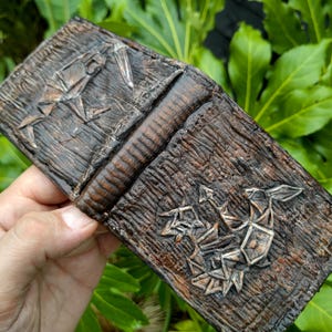 May include: A brown leather wallet with a geometric design embossed on the front. The design is made of silver metal and has a 3D effect.