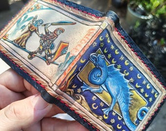 Puede incluir: Una cartera de cuero plegable con grabados y pinturas hechos a mano. El panel izquierdo muestra una figura con túnica beige y un guerrero con espada y escudo. El panel derecho presenta un dragón azul sobre un cielo nocturno con lunas.