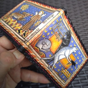 May include: A leather wallet featuring a detailed medieval-style illustration with a blue background, golden stars, and crescent moons. The artwork depicts a figure, a rabbit, a bird in a cage, and a cat, all framed by black stitched edges.