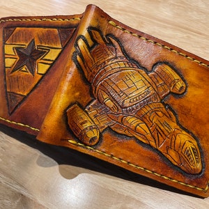May include: A brown leather wallet with a tooled design of a spaceship on the front. The wallet has a yellow stitching detail around the edges.