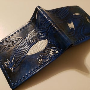 May include: A blue leather wallet with a white bird design embossed on the front. The wallet is folded in half and the stitching is visible.