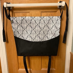 May include: A black and white patterned fabric baby carrier with black straps. The carrier is hanging on a white door handle.