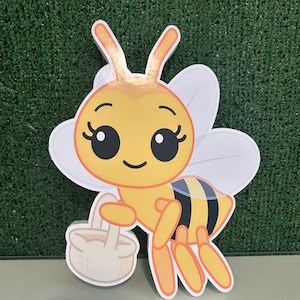 May include: A cartoon-style bee cutout with a yellow body, black and yellow striped abdomen, and white wings. The bee has large, friendly eyes and is holding a small basket. The antennae are gold. The cutout is likely for party decorations.