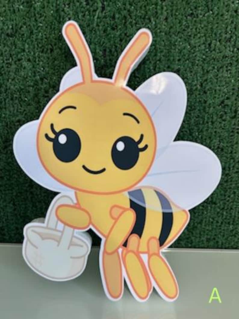 May include: A cartoon-style cutout of a bee, featuring a yellow body with black stripes and white wings. The bee has large, expressive eyes and is holding a small basket. The letter "A" is visible in the lower right corner.