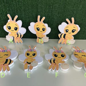 May include: A collection of seven bee-shaped cutouts. The bees are yellow and black with white wings, carrying baskets. Some bees have flower crowns. The cutouts are on a white surface with a green background.