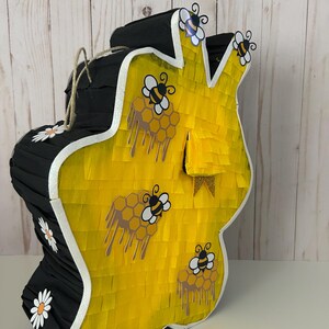 May include: A large, handmade pi&ntilde;ata in the shape of a honeycomb. The pi&ntilde;ata is primarily yellow with black accents and white trim. It features bee and honeycomb decorations, and a jute rope for hanging. Daisy accents are also present.