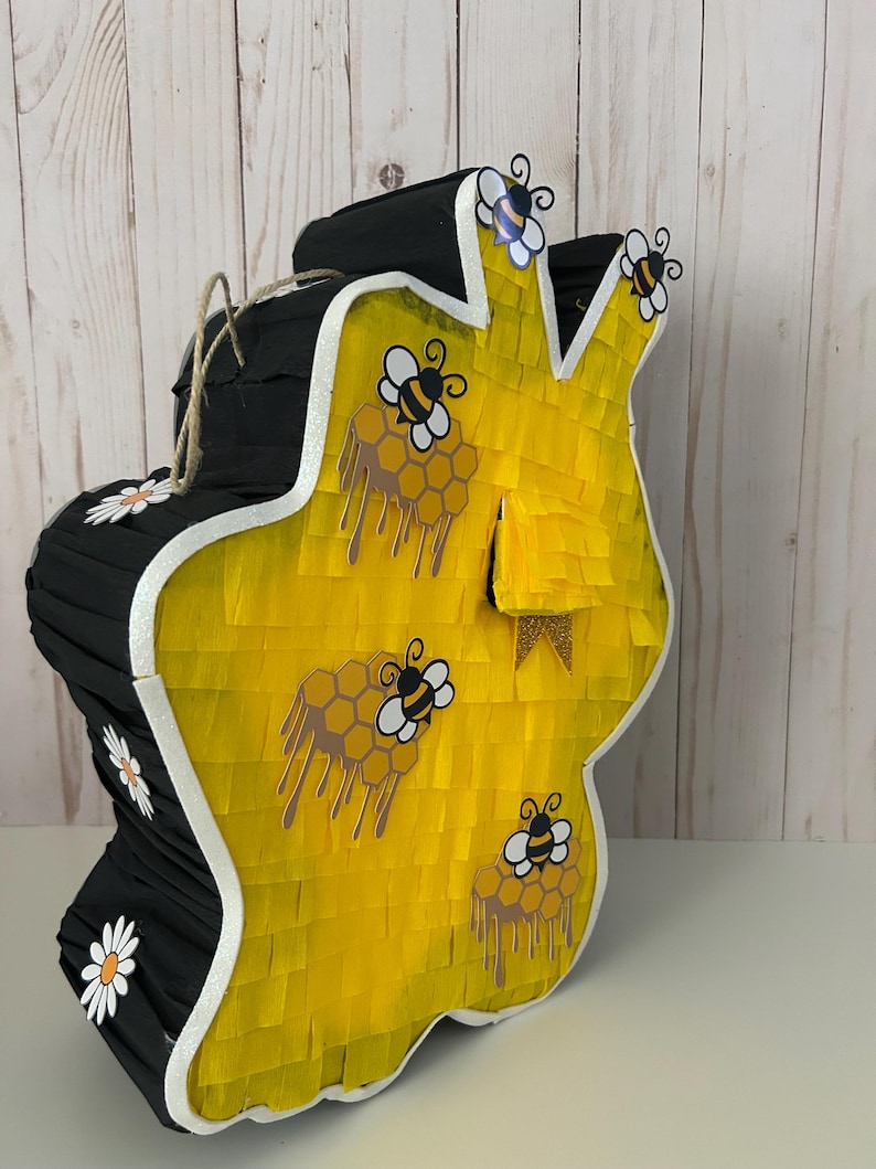 May include: A large, handmade pi&ntilde;ata in the shape of a honeycomb. The pi&ntilde;ata is primarily yellow with black accents and white trim. It features bee and honeycomb decorations, and a jute rope for hanging. Daisy accents are also present.